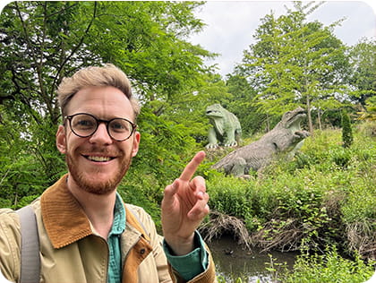 Closeup of a man standing in front of dinosaur statues in a green area of trees and shrubs